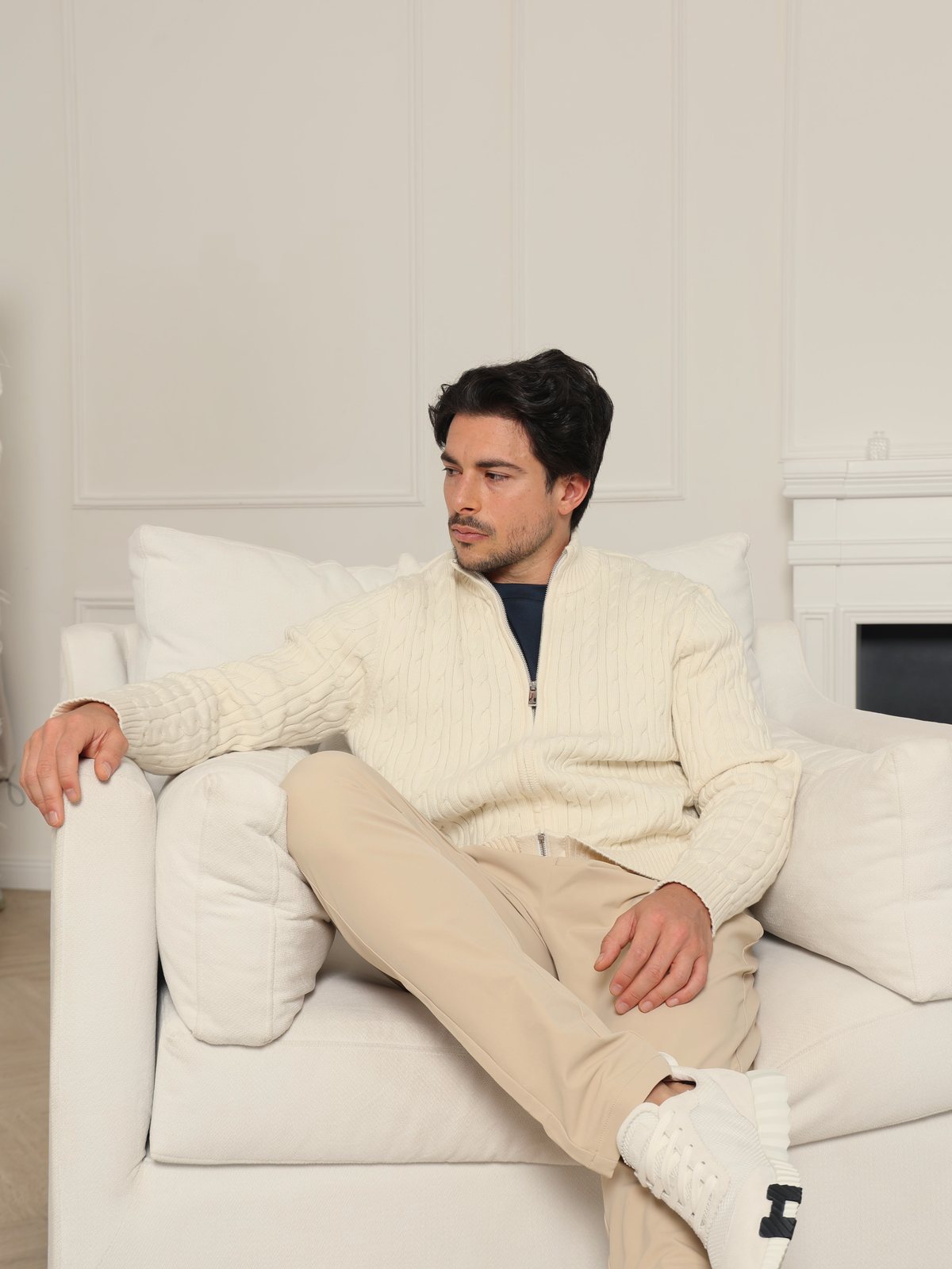 Man in an ivory cable-knit full zip on a white sofa in a panelled room